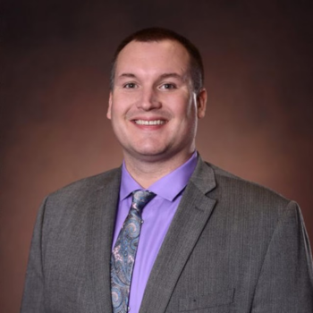 Man wearing suit, purple shirt and tie in front of reddish brown background 